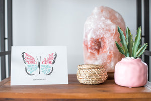 A card on a wood tabletop and on the right side of the card is a woven basket, a pink plant pot with a cactus in it and a pink crystal rock. The card features the words “Therefore if anyone is in Christ, the new creation has come. The old has gone. The new is here. 2 Corinthians 5:17.” The card design features an illustrated butterfly. 
