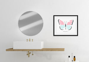 Artwork in a black frame featured in a bathroom next to a circle mirror and above a sink. The hand drawn artwork features the scripture verse "Therefore, if anyone is in Christ, the new creation has come. The old has gone. The new is here." The words are featured inside a butterfly. 