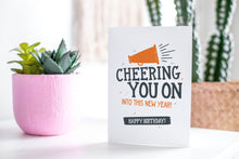 Load image into Gallery viewer, A greeting card featured standing up on a white tabletop with a pink plant pot in the background and some succulents in the pot. There’s a woven basket in the background with a cactus inside. The card features the words “Cheering You on into This New Year, Happy Birthday!”