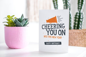 A greeting card featured standing up on a white tabletop with a pink plant pot in the background and some succulents in the pot. There’s a woven basket in the background with a cactus inside. The card features the words “Cheering You on into This New Year, Happy Birthday!”