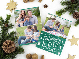 A photo of a two-sided Christmas card showing the front of the card on top of a brown wrapped gift on a white tabletop. Around the gift are pine needles, pinecones and wood star ornaments. The front of the card features a photo on the top and on the bottom is in green with words in white “Hallelujah Jesus is Here” with white stars around it. The back of the card features two photos. 