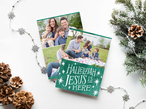 A photo of a double-sided Christmas card showing the front and back of the card laying on a white surface. Around the two sides of the card are pine cones, pine needles and a string of silver snowflake garland. The front of the card features a photo on the top and on the bottom is in green with words in white “Hallelujah Jesus is Here” with white stars around it. The back of the card features two photos.
