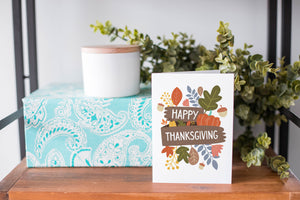 A greeting card is on a table top with a present in the background. There's greenery on top of the present. The card reads “Happy Thanksgiving” with illustrated leaves, a pumpkin and acorn.