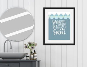 Artwork in a black frame featured in a bathroom next to a circle mirror and above a sink. The artwork features hand drawn lettering reading "When you go through deep waters, I will be with you."