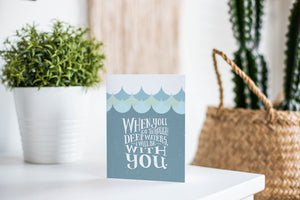 A greeting card is featured on a white tabletop with a white planter in the background with a green plant. There’s a woven basket in the background with a cactus inside. The card features the words “When you go through deep waters, I will be with you.”