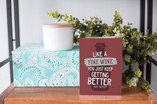 Load image into Gallery viewer, A greeting card is on a table top with a present in blue wrapping paper in the background. On top of the present is a candle and some greenery from a plant too. The card features the words “Like a Fine Wine, You Just Keep Getting Better, Happy Birthday.”