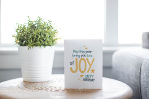 A card on a wood tabletop with an object in the background that is out of focus. The card features the words “May This Year Bring You Lots of Joy, Happy Birthday.”