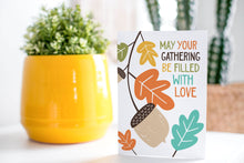 Load image into Gallery viewer, A greeting card is on a table top with a yellow plant pot and a green plant inside. The card features the words “May Your Gathering Be Filled with Love” with illustrated leaves and an acorn around the words.