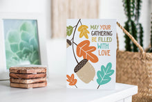 Load image into Gallery viewer, A greeting card featured standing up on a white tabletop with a framed photo of a succulent in the background and a stack of wooden coasters. There’s a woven basket in the background with a cactus inside. The card features the words “May Your Gathering Be Filled with Love” with illustrated leaves and an acorn around the words.