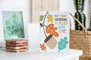 A greeting card featured standing up on a white tabletop with a framed photo of a succulent in the background and a stack of wooden coasters. There’s a woven basket in the background with a cactus inside. The card features the words “May Your Gathering Be Filled with Love” with illustrated leaves and an acorn around the words.