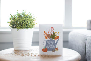 A greeting card is featured on round coffee table with a green plant and sofa in the background. The card features the words "May Your Table be Filled with Loved Ones" with the words inside an illustrated watering can with leaves coming out of the top.