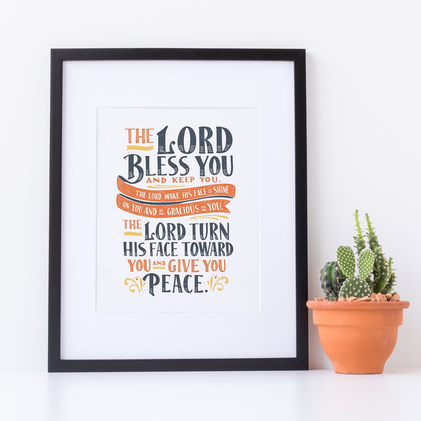 Artwork in a black frame with the with a white matte. The frame is leaning on a white counter with a terracota pot with a catcus next to it. The artwork features hand drawn lettering of the Bible verse Numbers 6:24-26 reading 