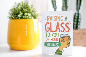 A greeting card is on a table top with a yellow plant pot and a green plant inside. The card features the words “Raising a glass to you on your birthday” with an illustrated hand raising a glass.