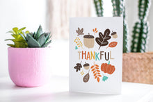 Load image into Gallery viewer, A greeting card featured standing up on a white tabletop with a pink plant pot with succulents. There’s a woven basket in the background with a cactus inside. The card features the words “Thankful&quot; with illustrated leaves and an acorn around the word.