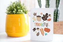 Load image into Gallery viewer, A greeting card is on a table top with a yellow plant pot and a green plant inside. The card features the words “Thankful&quot; with illustrated leaves and an acorn around the word.