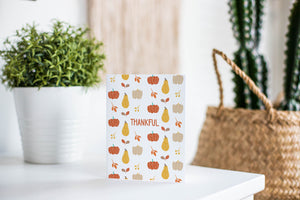 A greeting card featured standing up on a white tabletop with a white plant pot with a green plant. There’s a woven basket in the background with a cactus inside. The card features the word "Thankful" with a pattern of illustrated pumpkins and leaves behind the word. 