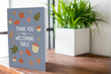 Load image into Gallery viewer, A photo of a card featured on a tabletop next to a white planter filled with a green plant. ​​The card features the words &quot;Thank You for a Welcoming Table&quot; with illustrated leaves and acorns around the words.