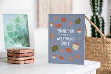 Load image into Gallery viewer, A greeting card featured standing up on a white tabletop with a stack of coasters next to it. There’s a woven basket in the background with a cactus inside. The card features the words &quot;Thank You for a Welcoming Table&quot; with illustrated leaves and acorns around the words. 