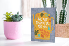 Load image into Gallery viewer, A greeting card featured standing up on a white tabletop with a pink plant pot with succulents. The card features the words &quot;We are Thankful for You&quot; with the words featured inside an illustrated teapot. 