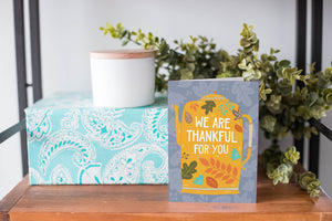 A greeting card is on a table top with a present in the background. There's greenery on top of the present. The card features the words "We are Thankful for You" with the words featured inside an illustrated teapot. 
