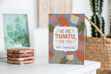 Load image into Gallery viewer, A greeting card featured standing up on a white tabletop with a stack of coasters next to it. There’s a woven basket in the background with a cactus inside. The card features the words &quot;We are Thankful for You, Happy Thanksgiving&quot; with illustrated leaves and acorns around the words. 