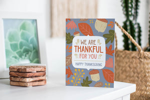 A greeting card featured standing up on a white tabletop with a stack of coasters next to it. There’s a woven basket in the background with a cactus inside. The card features the words "We are Thankful for You, Happy Thanksgiving" with illustrated leaves and acorns around the words. 