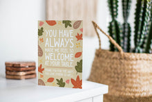 Load image into Gallery viewer, A greeting card featured standing up on a white tabletop with a stack of coasters next to it. There’s a woven basket in the background with a cactus inside. The card features the words &quot;You have always made me feel so welcome at your table. Thank you for the ways You love others so well&quot; with illustrated leaves surrounding the words. 
