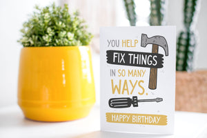 A greeting card is on a table top with a yellow plant pot and a green plant inside. The card features the words “You help fix things in so many ways. Happy Birthday” featuring an illustrated hammer.