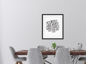 Artwork featured on a wall in a dining room with a dark wood table and grey dining chairs. The artwork features hand drawn lettering reading "Act Justly, Love Mercy, Walk Humbly with your God" - Micah 6:8.