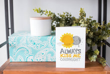 Load image into Gallery viewer, A greeting card is on a table top with a present in blue wrapping paper in the background. On top of the present is a candle and some greenery from a plant too. The card features the words “Always kiss me goodnight” with an illustrated sun and moon giving each other a kiss.
