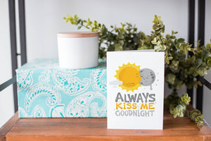 A greeting card is on a table top with a present in blue wrapping paper in the background. On top of the present is a candle and some greenery from a plant too. The card features the words “Always kiss me goodnight” with an illustrated sun and moon giving each other a kiss.