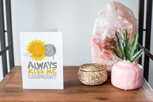 Load image into Gallery viewer, A card on a wood tabletop and on the right side of the card is a woven basket, a pink plant pot with a cactus in it and a pink crystal rock. The card features the words “Always kiss me goodnight” with an illustrated sun and moon giving each other a kiss.