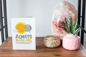 A card on a wood tabletop and on the right side of the card is a woven basket, a pink plant pot with a cactus in it and a pink crystal rock. The card features the words “Always kiss me goodnight” with an illustrated sun and moon giving each other a kiss.