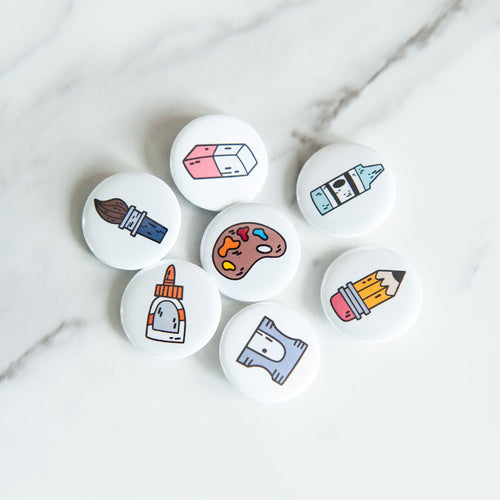 A set of seven button pins on a white tabletop. The pins feature illustrated designs of a paintbrush, eraser, crayon, pencil, paint palette, pencil sharpener, and a glue bottle. 