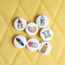 Load image into Gallery viewer, A set of seven button pins on a yellow background. The pins feature illustrated designs of a paintbrush, eraser, crayon, pencil, paint palette, pencil sharpener, and a glue bottle. 