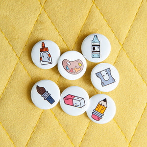 A set of seven button pins on a yellow background. The pins feature illustrated designs of a paintbrush, eraser, crayon, pencil, paint palette, pencil sharpener, and a glue bottle. 