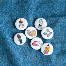 Load image into Gallery viewer, A set of seven button pins on a jean jacket. The pins feature illustrated designs of a paintbrush, eraser, crayon, pencil, paint palette, pencil sharpener, and a glue bottle. 