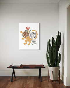 A white canvas with artwork hanging on a wall above a bench in an entryway. The artwork features an illustrated lion with a crown and reads "Course He Isn't Safe, But He's Good. He's the King."