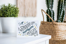 Load image into Gallery viewer, A greeting card is featured on a white tabletop with a white planter in the background with a green plant. There’s a woven basket in the background with a cactus inside. The card features the words “A smooth sea never made a skilled sailor.”
