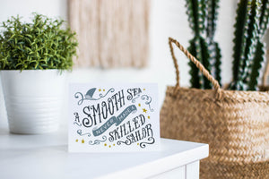 A greeting card is featured on a white tabletop with a white planter in the background with a green plant. There’s a woven basket in the background with a cactus inside. The card features the words “A smooth sea never made a skilled sailor.”