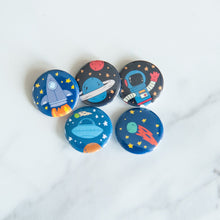 Load image into Gallery viewer, A set of five buttons on a white tabletop. The buttons feature space-themed illustrations of planets, a spaceship, a rocket and an astronaut. 