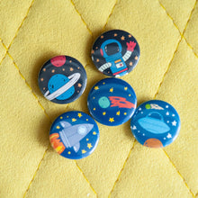 Load image into Gallery viewer, A set of five buttons on a yellow background. The buttons feature space-themed illustrations of planets, a spaceship, a rocket and an astronaut. 