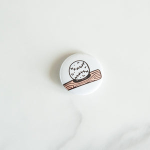 A buttons on a white tabletop. The button features an illustrations of a baseball with baseball bat. 