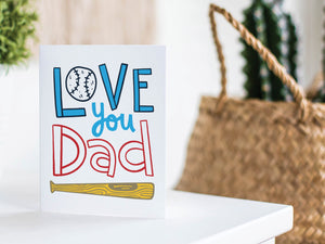 A greeting card is featured on a white tabletop with a white planter in the background with a green plant. There’s a woven basket in the background with a cactus inside. The card features the words “Love You Dad” with an illustrated baseball as the “O” of love and a baseball bat featured at the bottom of the words. 