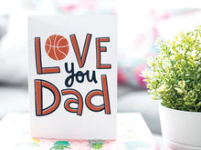 Load image into Gallery viewer, A greeting card is on a table top with a gift in pink wrapping paper. Next to the gift is a white plant pot with a green plant. The card features the words “Love you Dad” with an illustrated basketball as the “O” of love. 