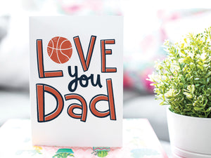 A greeting card is on a table top with a gift in pink wrapping paper. Next to the gift is a white plant pot with a green plant. The card features the words “Love you Dad” with an illustrated basketball as the “O” of love. 