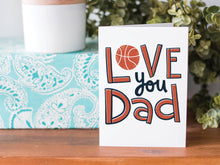 Load image into Gallery viewer, A greeting card is on a table top with a present in blue wrapping paper in the background. On top of the present is a candle and some greenery from a plant too. The card features the words  “Love you Dad” with an illustrated basketball as the “O” of love. 