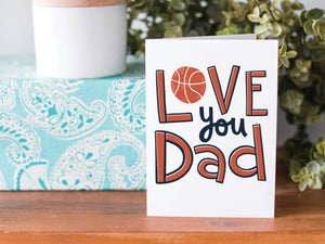A greeting card is on a table top with a present in blue wrapping paper in the background. On top of the present is a candle and some greenery from a plant too. The card features the words  “Love you Dad” with an illustrated basketball as the “O” of love. 