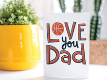 Load image into Gallery viewer, A greeting card is on a table top with a yellow plant pot and a green plant inside. The card features the words “Love you Dad” with an illustrated basketball as the “O” of love. 