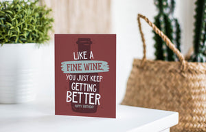 A greeting card is featured on a white tabletop with a white planter in the background with a green plant. There’s a woven basket in the background with a cactus inside. The card features the words “Like a Fine Wine, You Just Keep Getting Better, Happy Birthday.”